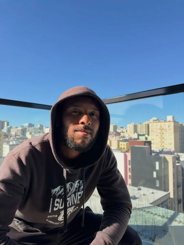 Chantz Rivera poses outdoors against a city skyline backdrop after walking 2,500 miles from New York to San Francisco.