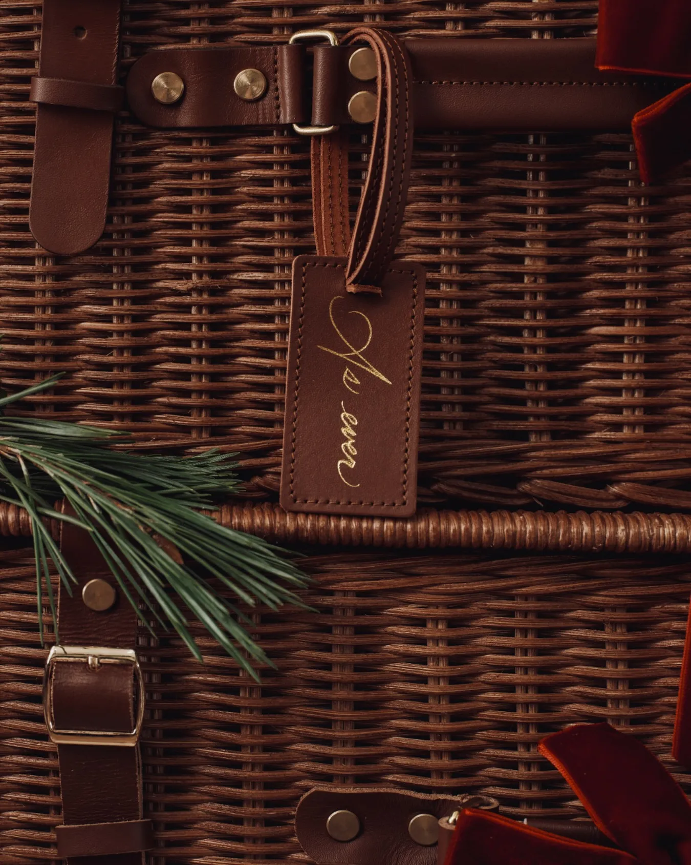 A brown wicker hamper with leather straps, a luggage tag, and a pine branch.