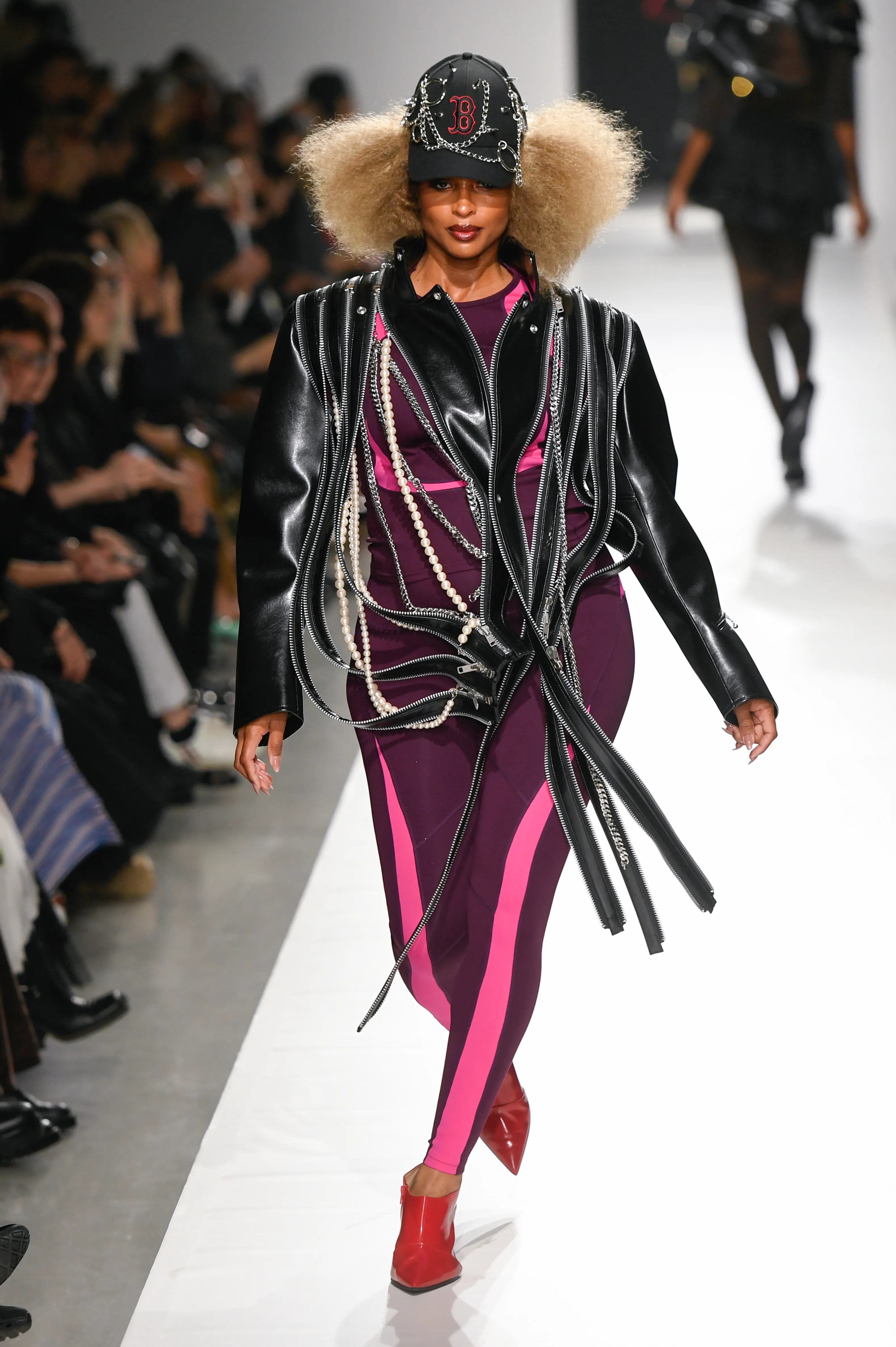 Ciara walks the runway at the Junya Watanabe fashion show wearing a purple bodysuit with pink stripes and a black leather jacket embellished with zippers, chains, and pearls, paired with a beaded cap and red shoes.