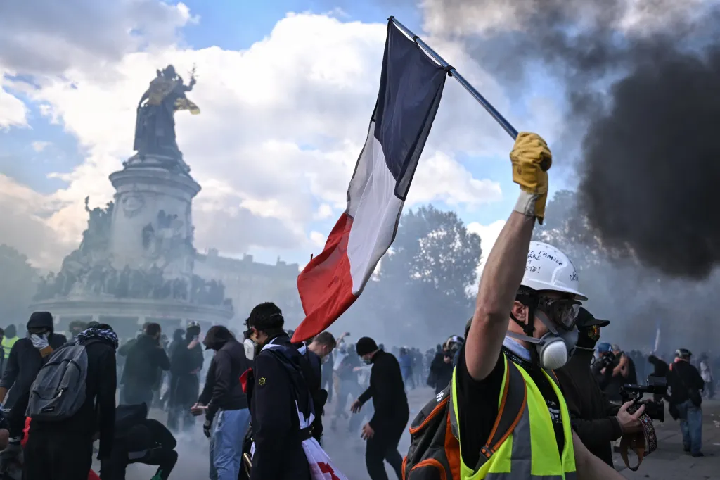 Protestor raises a French flag during mass demonstrations in Paris, with a statue and smoke in the background.