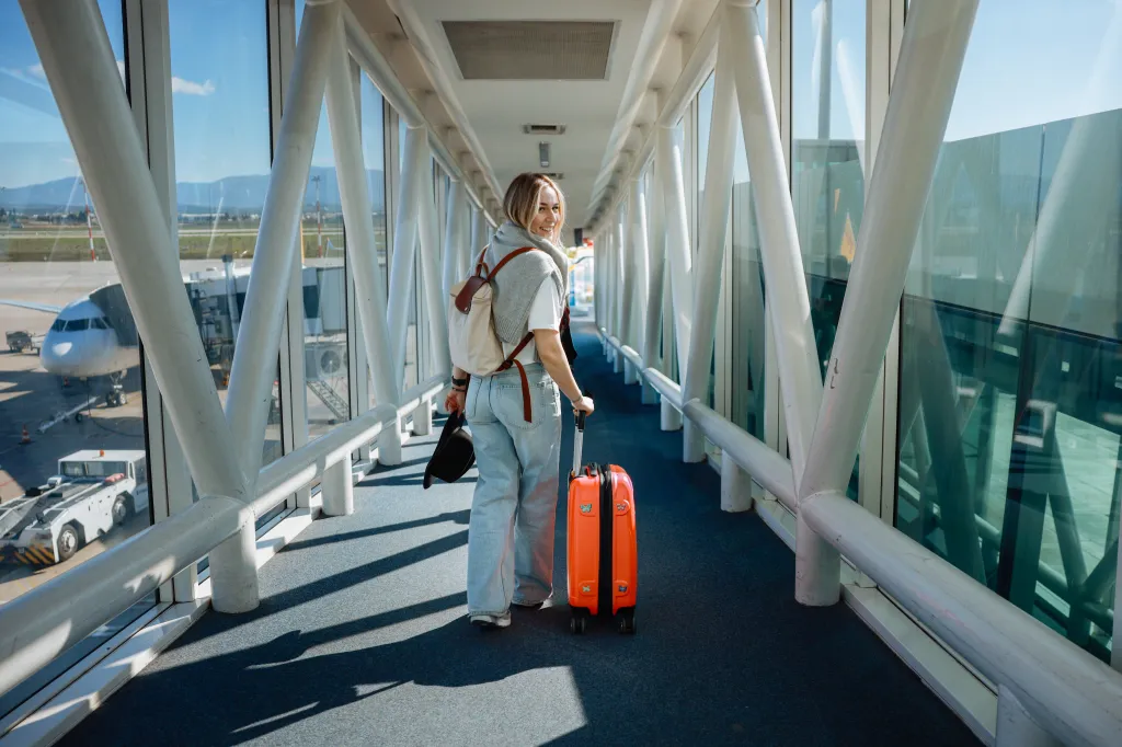 A female traveler in casual clothing pulls a wheelie suitcase through an airport jetway.