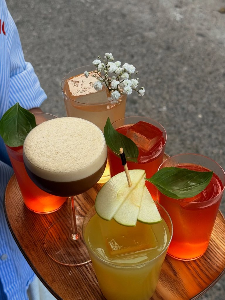 A tray holding a variety of cocktails, including a creamy espresso martini, yellow juice garnished with apple slices, and several reddish drinks adorned with fresh herbs and flowers.