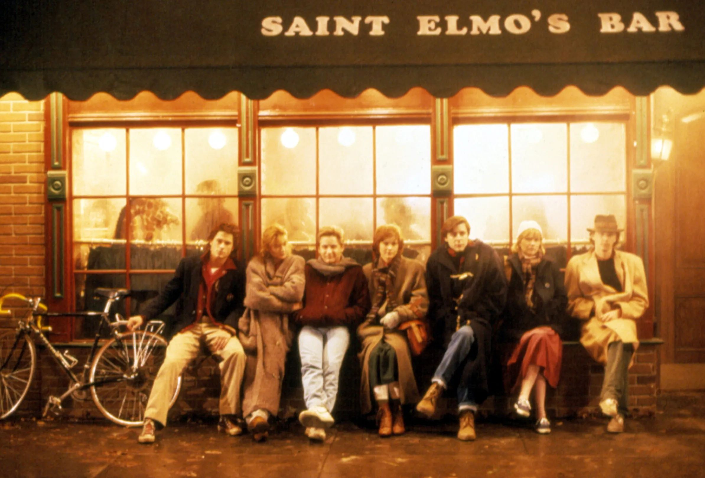 A group shot of Rob Lowe, Demi Moore, Emilio Estevez, Ally Sheedy, Judd Nelson, Mare Winningham, and Andrew McCarthy sitting in front of Saint Elmo's Bar.