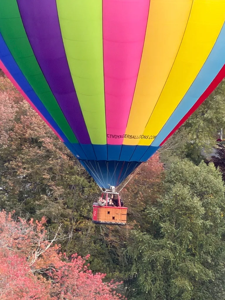 A brightly colored, multi-striped hot air balloon flies low over autumn trees, with passengers visible in the basket.