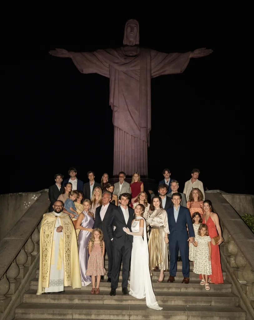 Conor Kennedy and his bride pose with wedding guests on steps in front of the illuminated Christ the Redeemer statue at night.