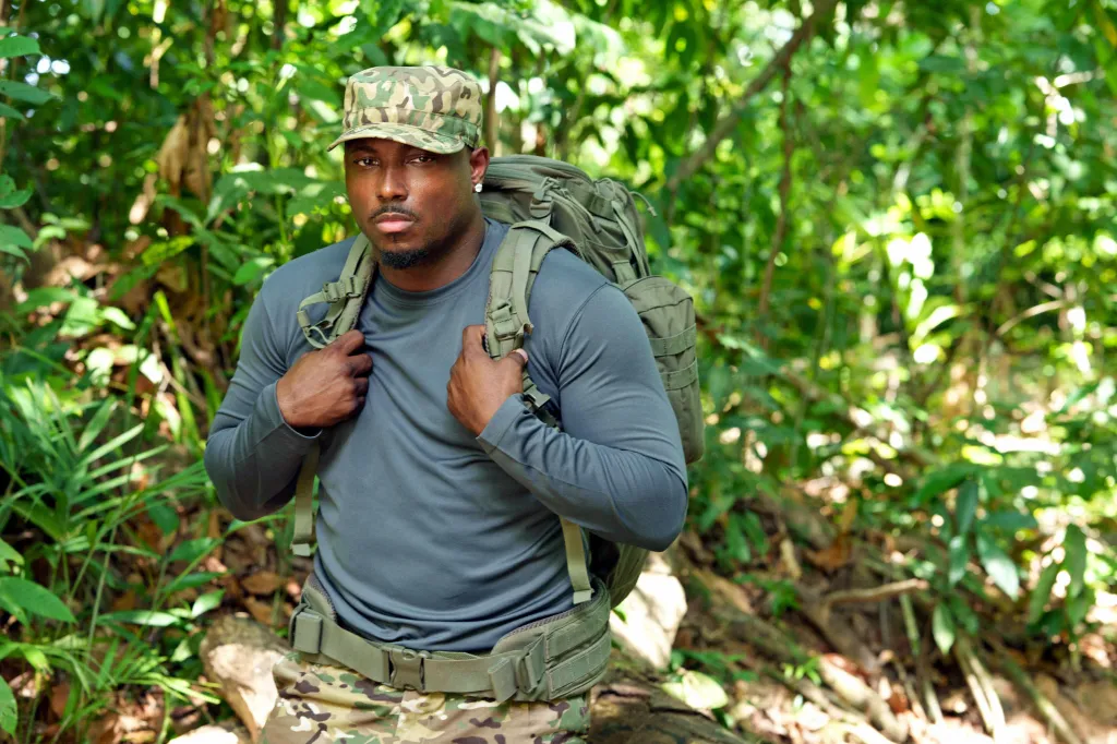 LeSean McCoy wearing a camouflage cap, grey long-sleeve shirt, and carrying a large backpack in a jungle setting.