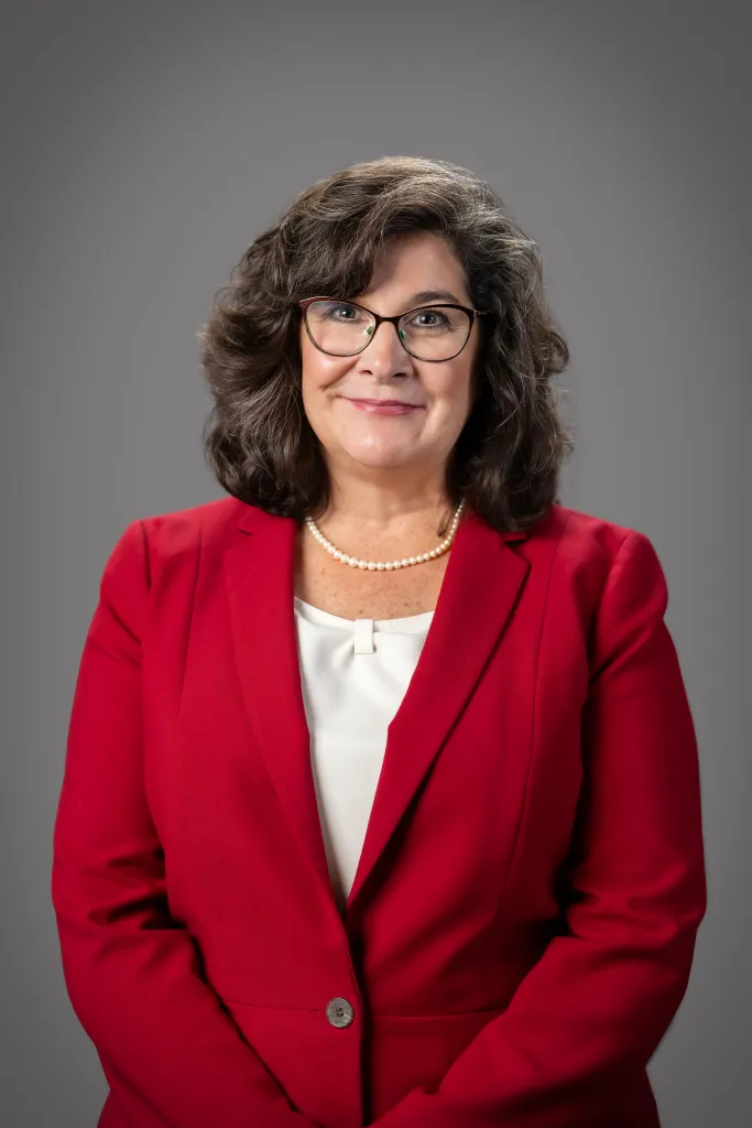 Portrait of Kim Geyer wearing a red blazer and glasses against a gray background.