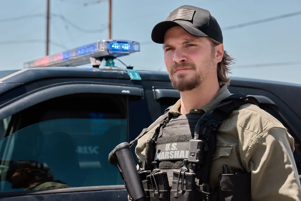 Luke Grimes as U.S. Marshal Kayce Dutton in front of a police vehicle with flashing lights.