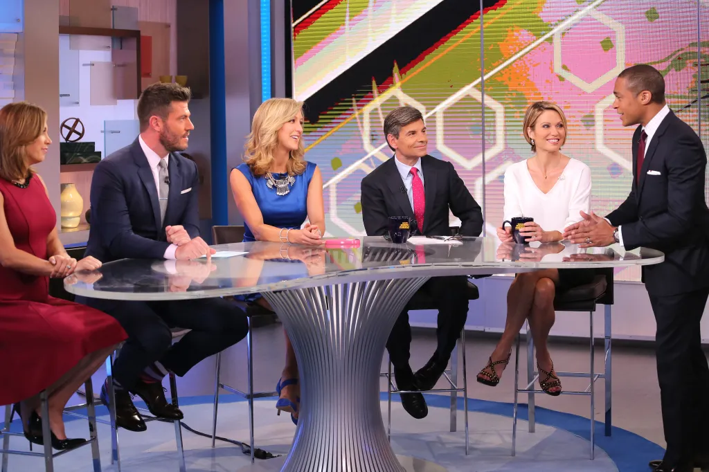 The anchors of Good Morning America are seated around a glass desk, engaged in a discussion.
