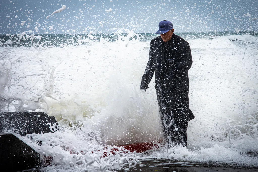 A man in a trench coat and cap braves large tidal swells crashing against the Southern California coastline.