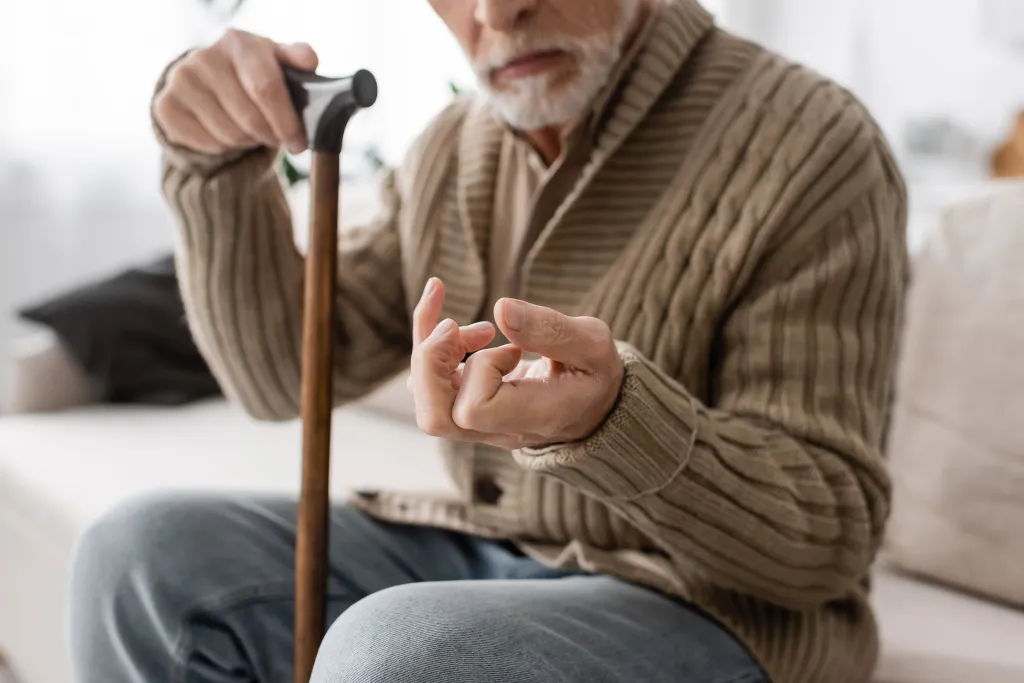 A senior man with Parkinson's disease sitting on a couch at home, holding a walking cane and exhibiting hand tremors.
