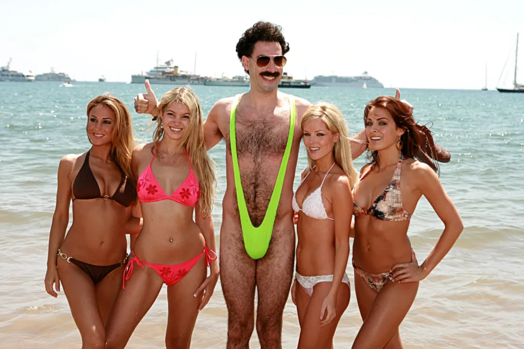 Sacha Baron Cohen as Borat, wearing a neon green mankini and sunglasses, standing on a beach with four women in bikinis.