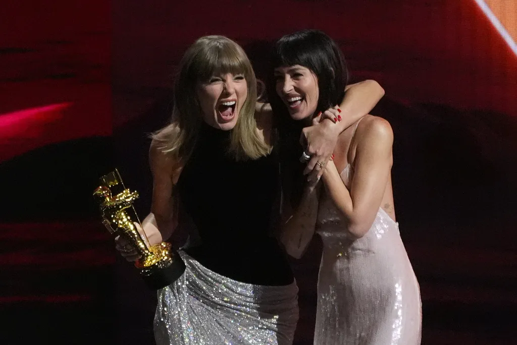 Taylor Swift and Dakota Johnson embrace and smile, with Swift holding a golden trophy.