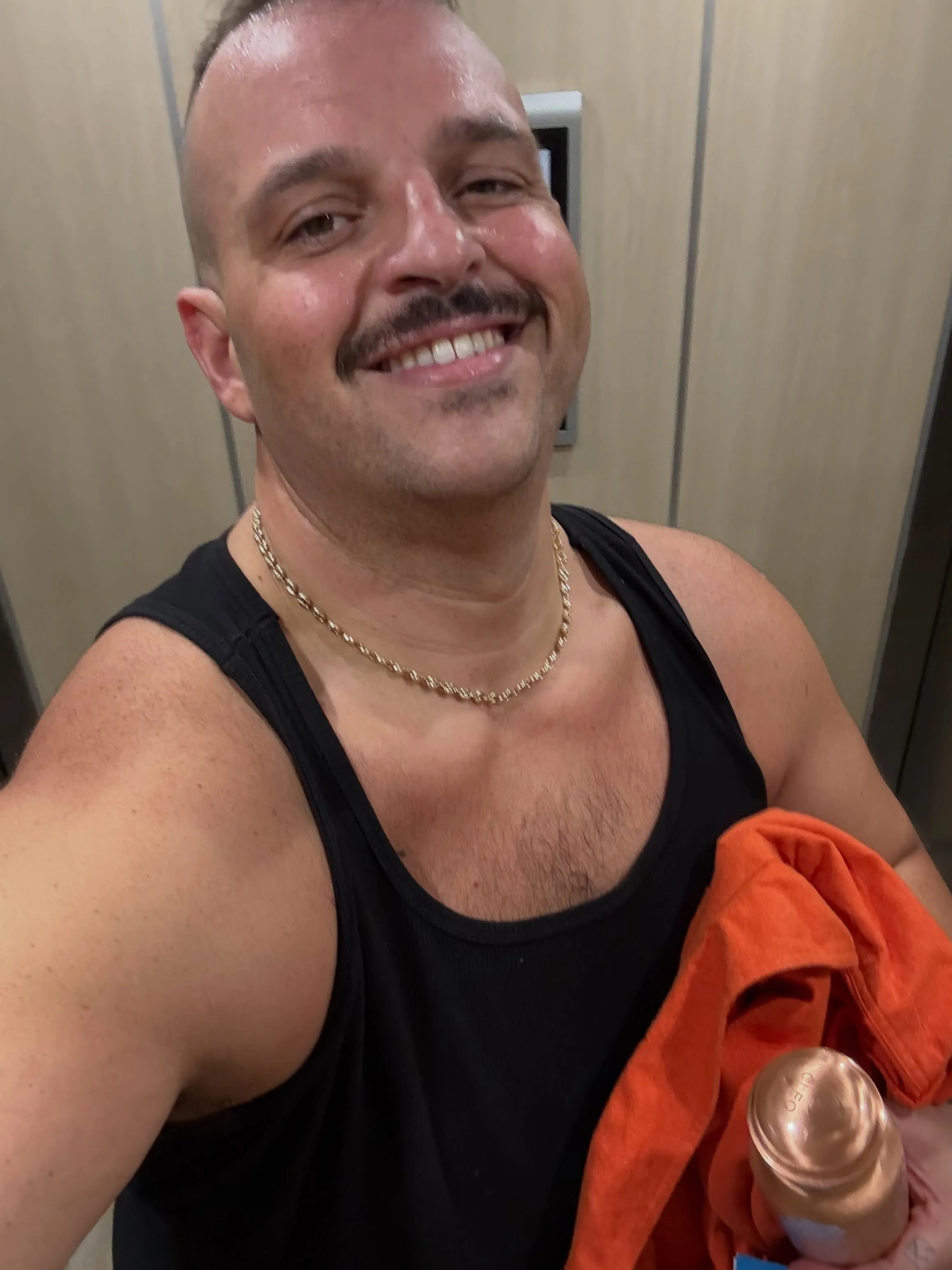 Daniel Franzese smiling in a black tank top, holding an orange garment and a bottle.