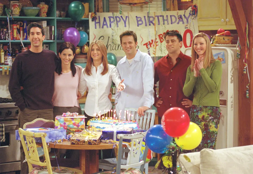 The cast of Friends standing behind a birthday table with balloons, a cake, and presents.
