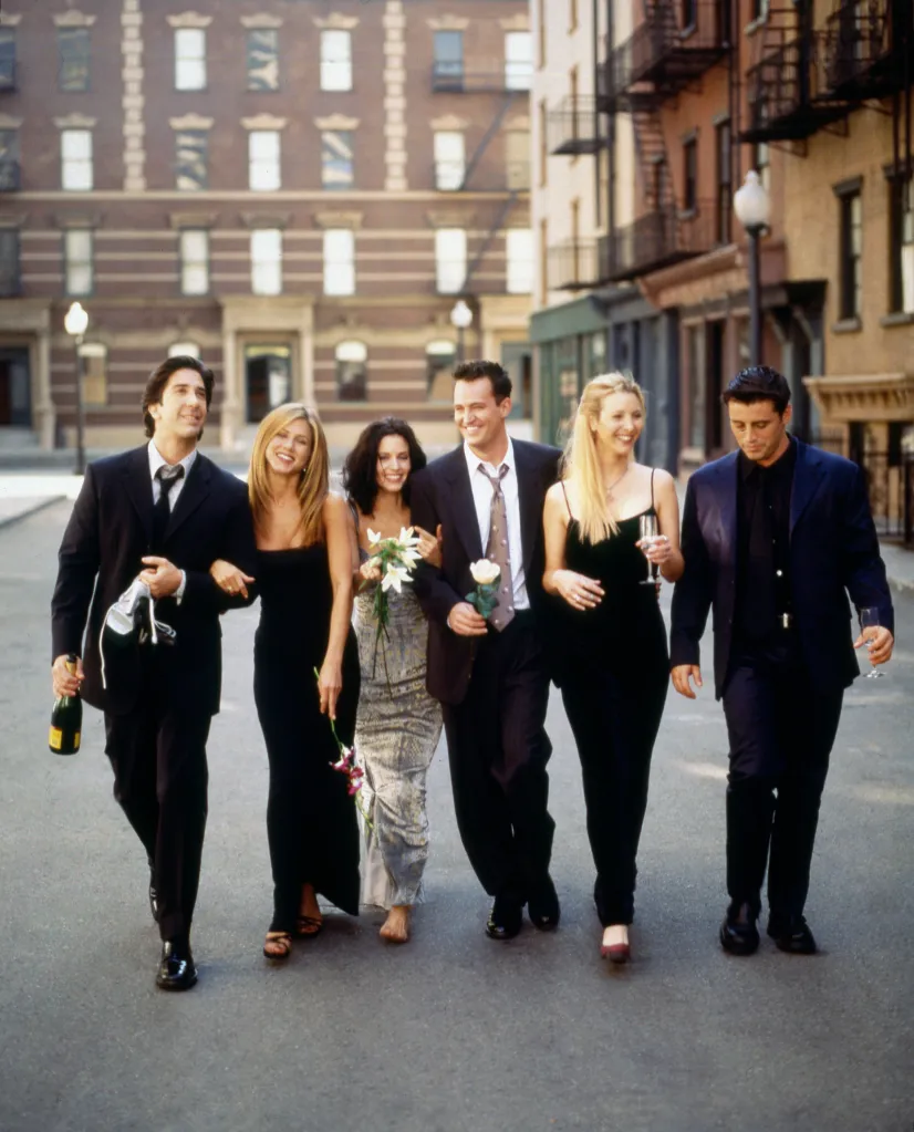 The cast of Friends walking down a city street in formal wear, carrying champagne and flowers.