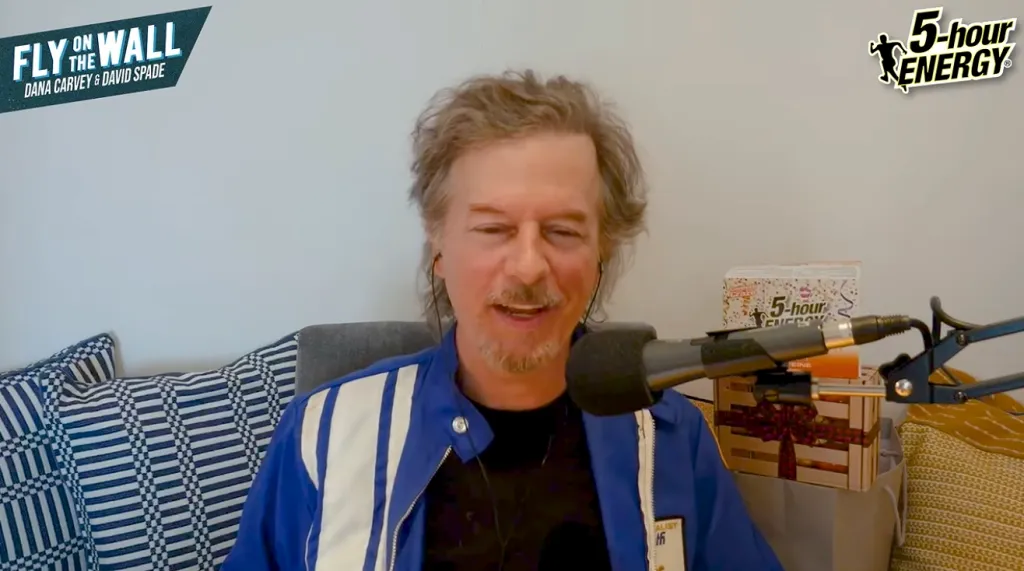 David Spade smiling while sitting on a couch with a microphone in front of him.