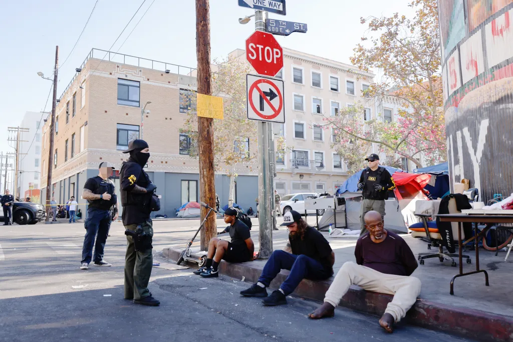 Law enforcement officers standing guard over several individuals sitting with their hands behind their backs on a curb in Skid Row during Operation Free Skid Row.