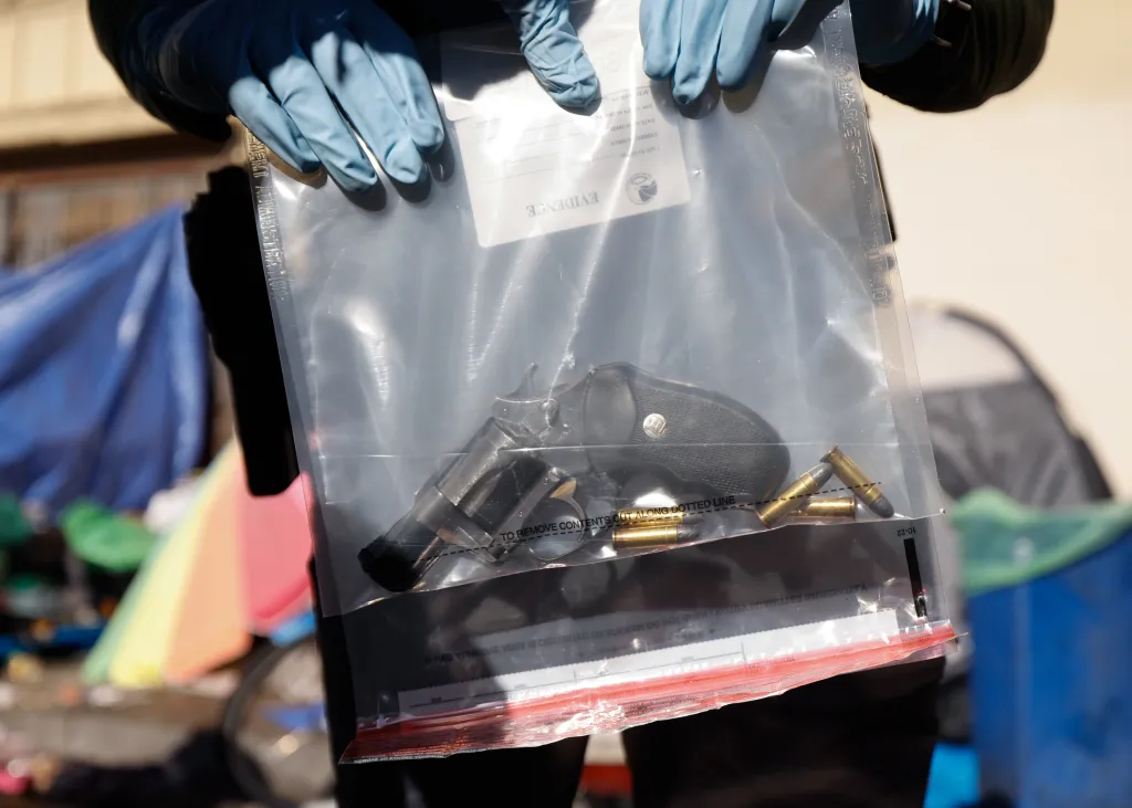 An officer wearing blue gloves holds up an evidence bag containing a revolver and several rounds of ammunition in Skid Row.