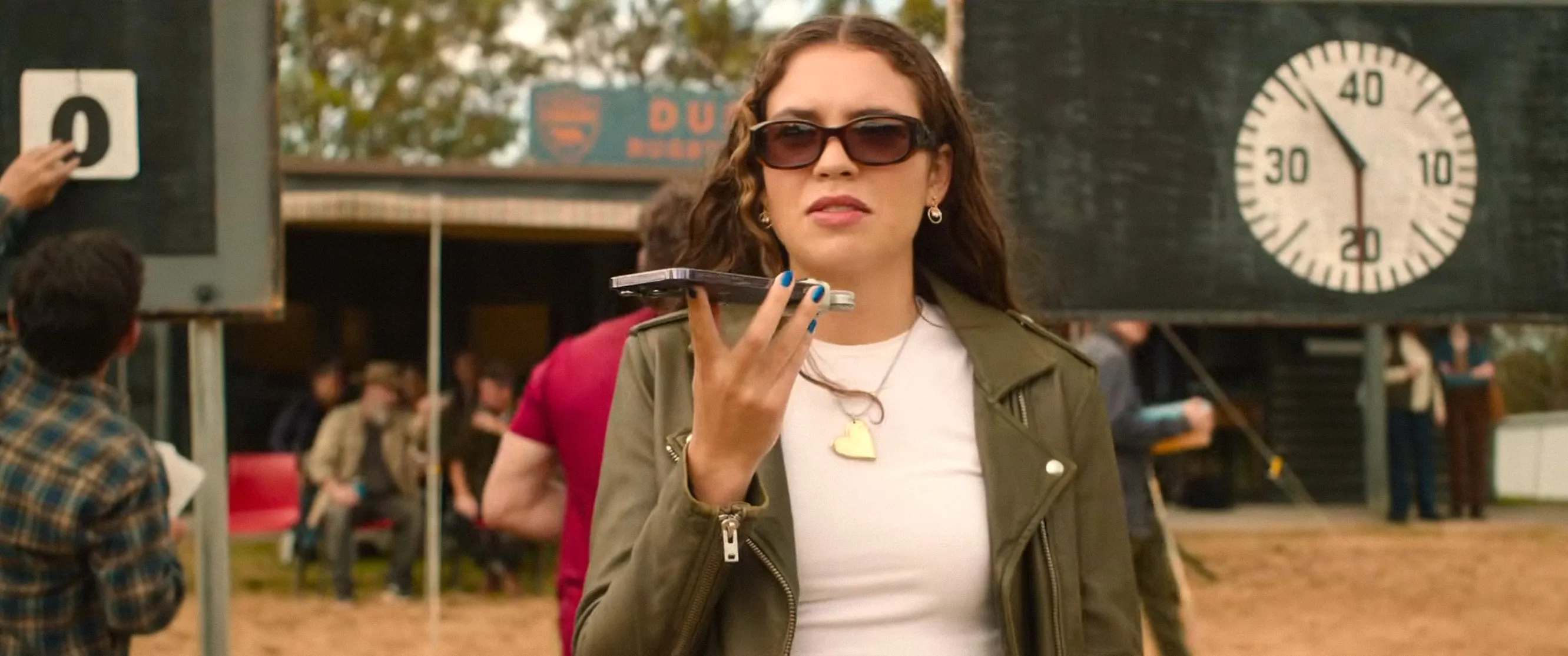 Charlotte MacInnes wearing sunglasses and holding a phone on speaker, in a scene from the film The Deb.