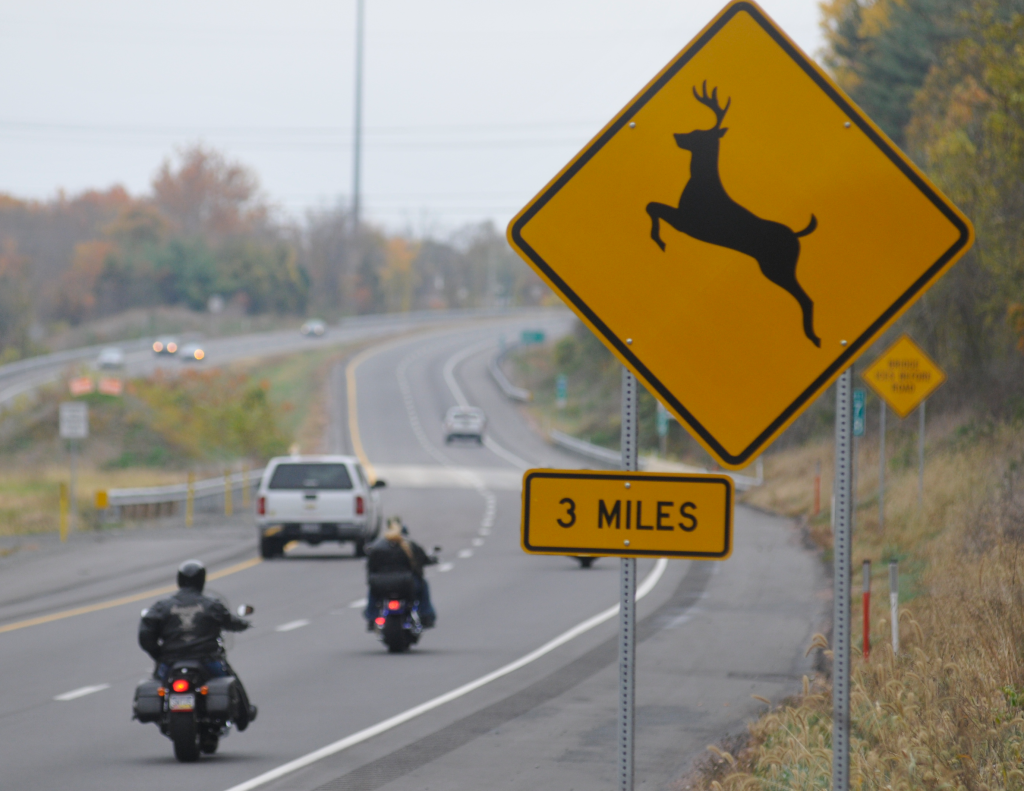 A highway with cars and motorcycles with a deer crossing sign
