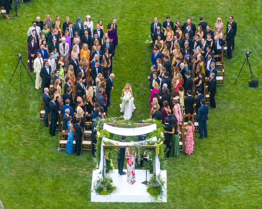 An aerial shot shows a bride walking down the grassy aisle of an outdoor wedding ceremony, with rows of seated guests forming the shape of a heart around the white chuppah.
