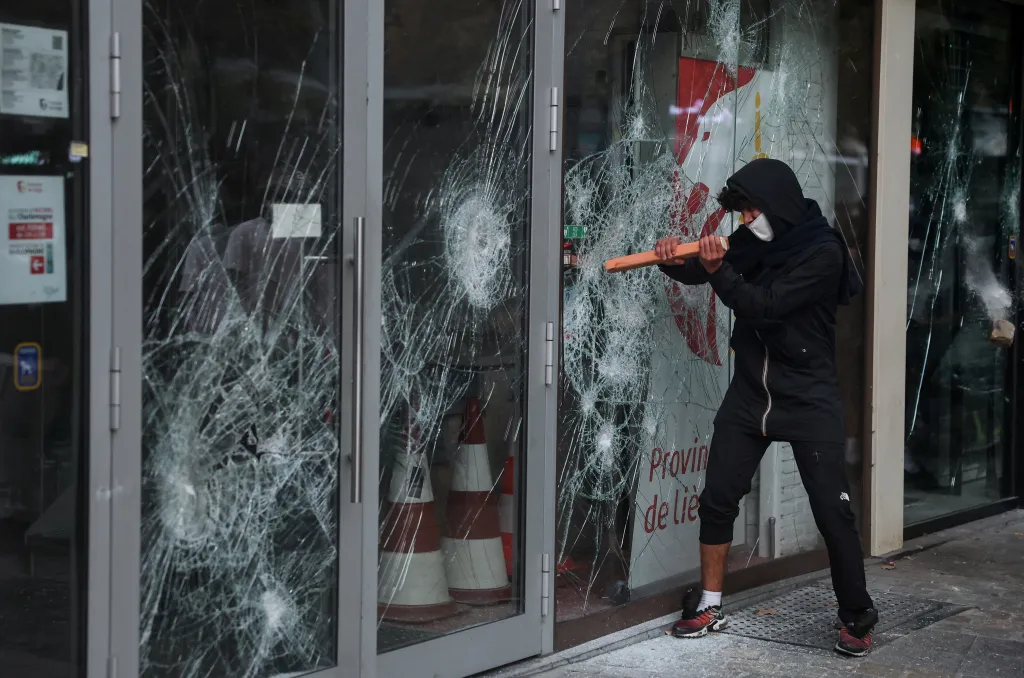 A demonstrator in a black hoodie and mask breaks the glass of a storefront during a protest in Liege.