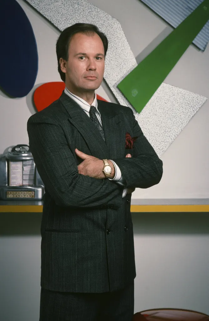 Dennis Haskins as Mr. Richard Belding from Saved by the Bell, standing with arms crossed, wearing a striped suit and tie.