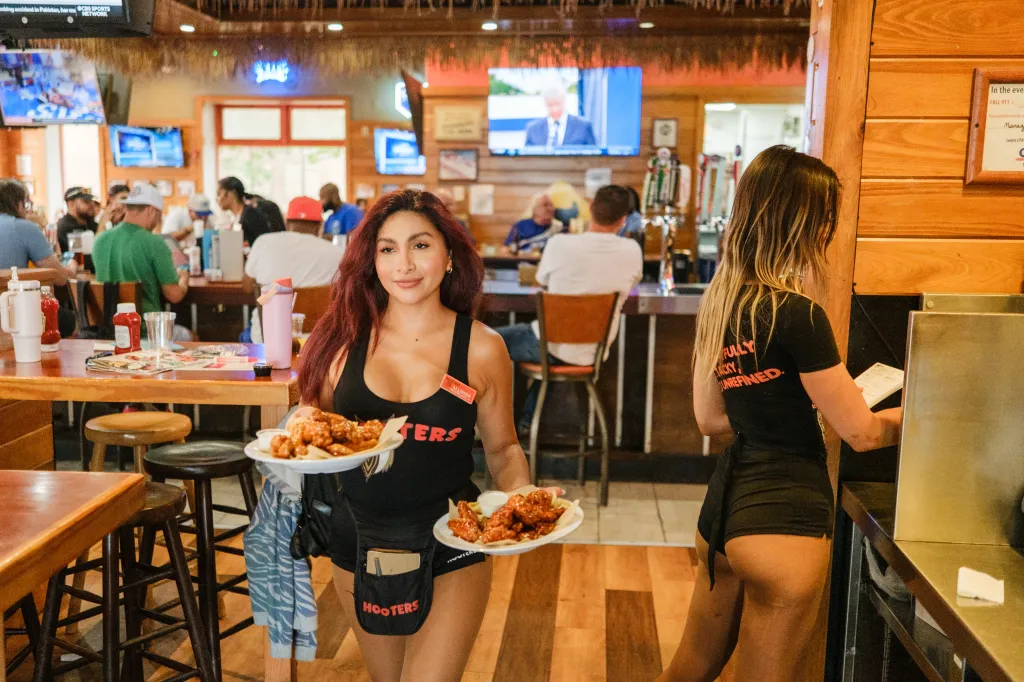 Inside a busy Hooters restaurant, a waitress holds two plates of chicken wings while looking at the camera, with another waitress standing nearby.