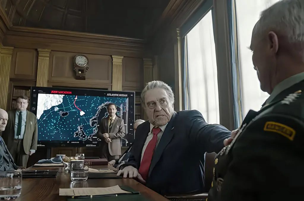 John Goodman in a suit sits at a table and gestures toward a military officer, with other men and a large map screen in the background.