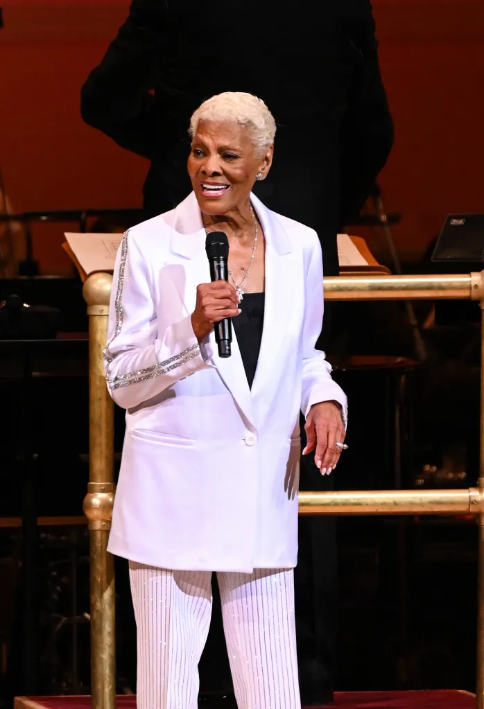 Dionne Warwick performs onstage wearing a white suit and holding a microphone.