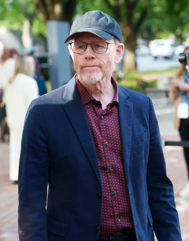 Director Ron Howard wearing a navy blazer, patterned shirt, cap, and glasses.