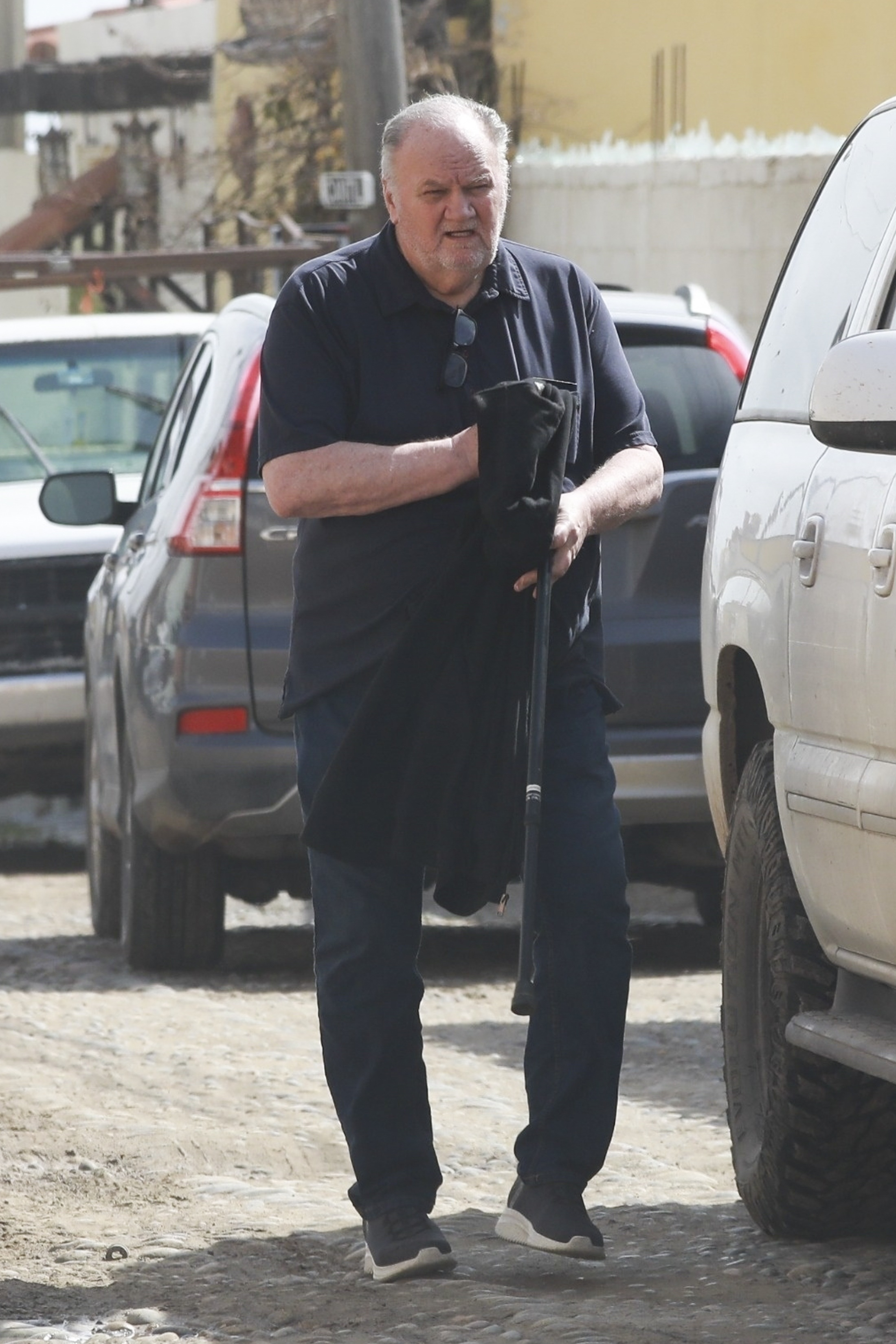 Thomas Markle walks down a cobbled street in Mexico, holding a jacket and a cane between parked cars.