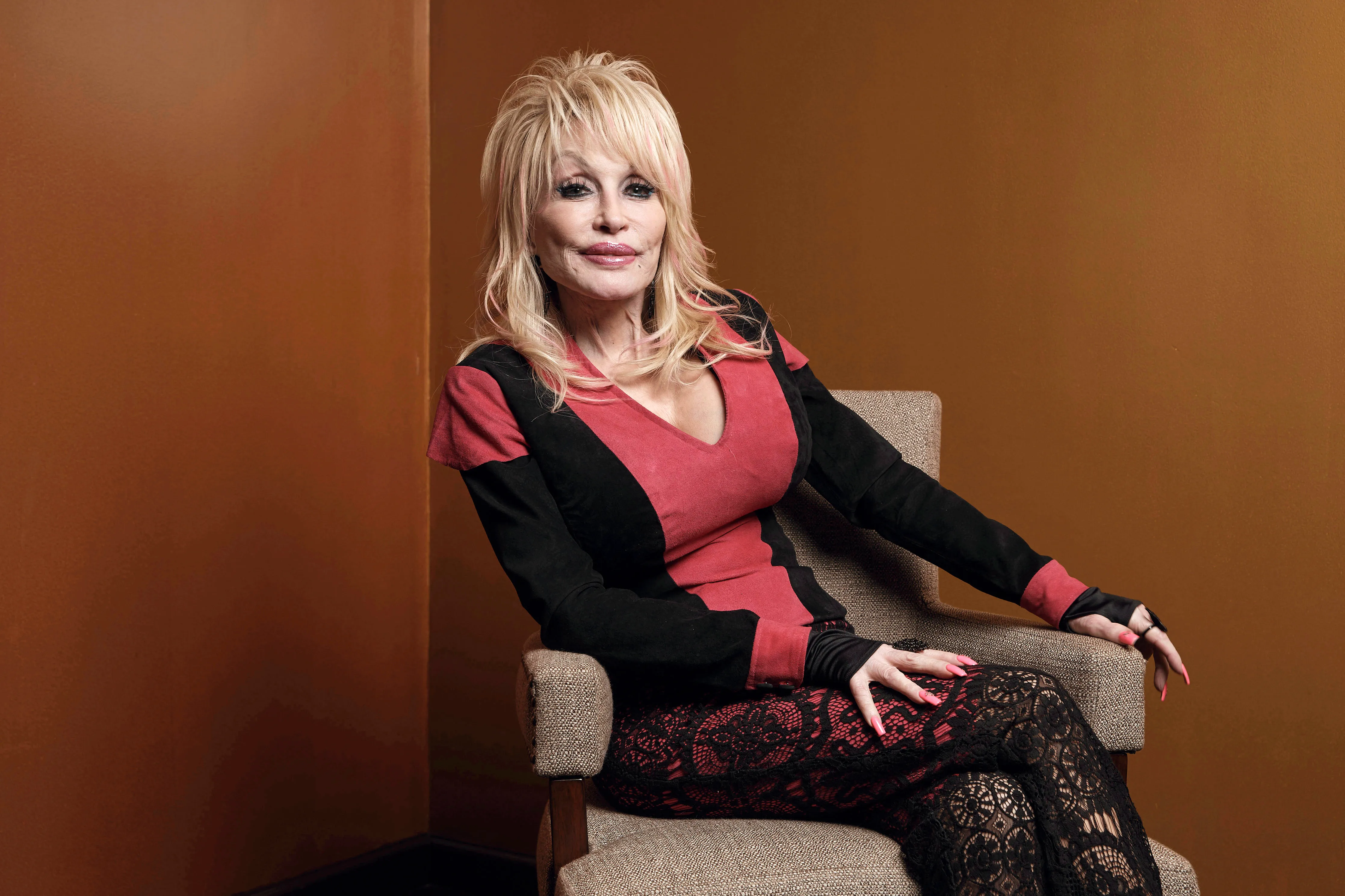 Dolly Parton poses seated in a chair against a brown backdrop.