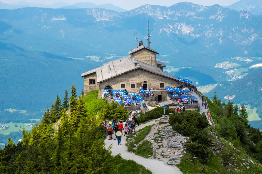 A general view of the Kehlsteinhaus, also known as The Eagle's Nest, perched on a mountain peak with visitors walking along the path and sitting at outdoor tables under blue umbrellas.