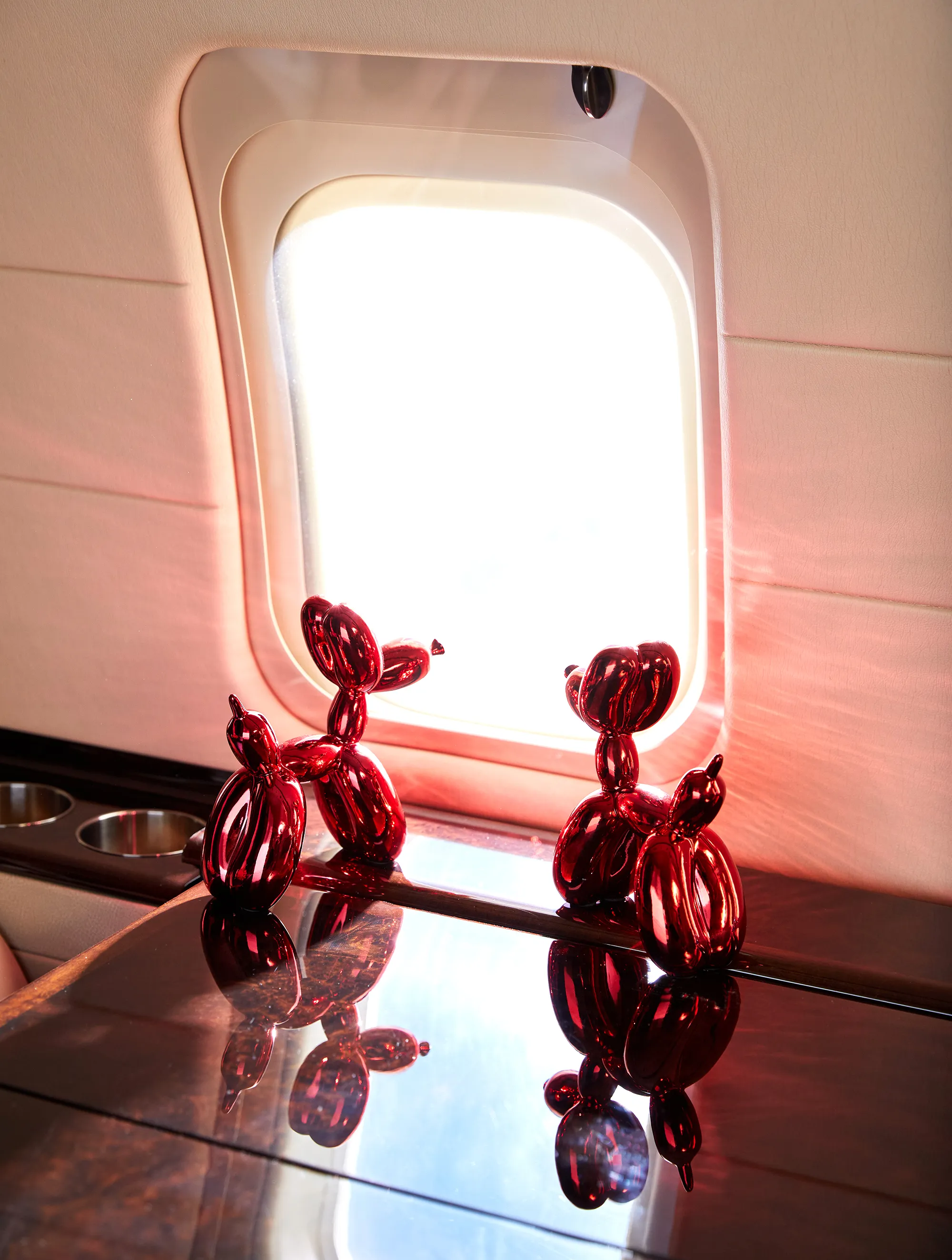 Two red balloon dog sculptures on a glossy surface near an airplane window on the BLADEone jet.