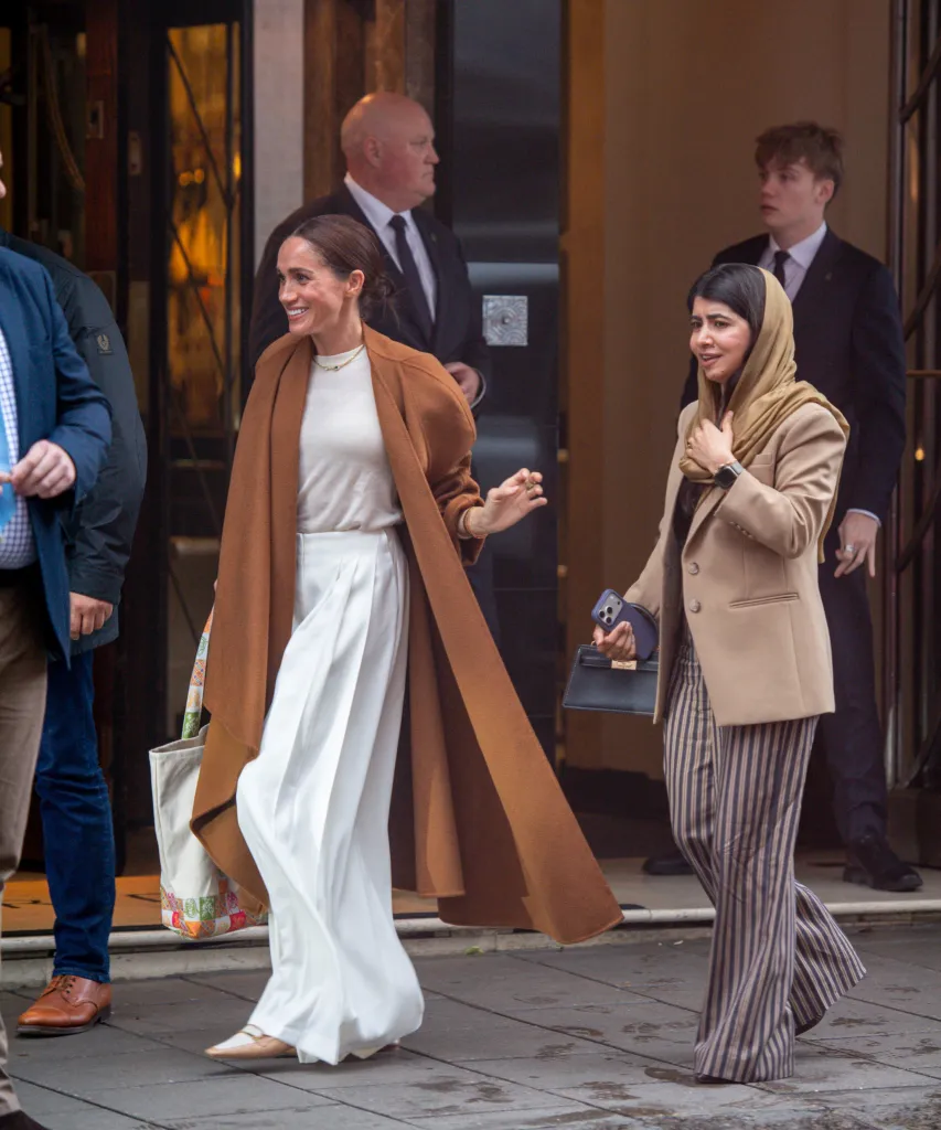 Meghan Markle, in a brown coat, cream top and white pants, and Malala Yousafzai, in a brown jacket, brown headscarf and brown striped pants, eaving Claridge’s hotel in London, accompanied by security personnel.