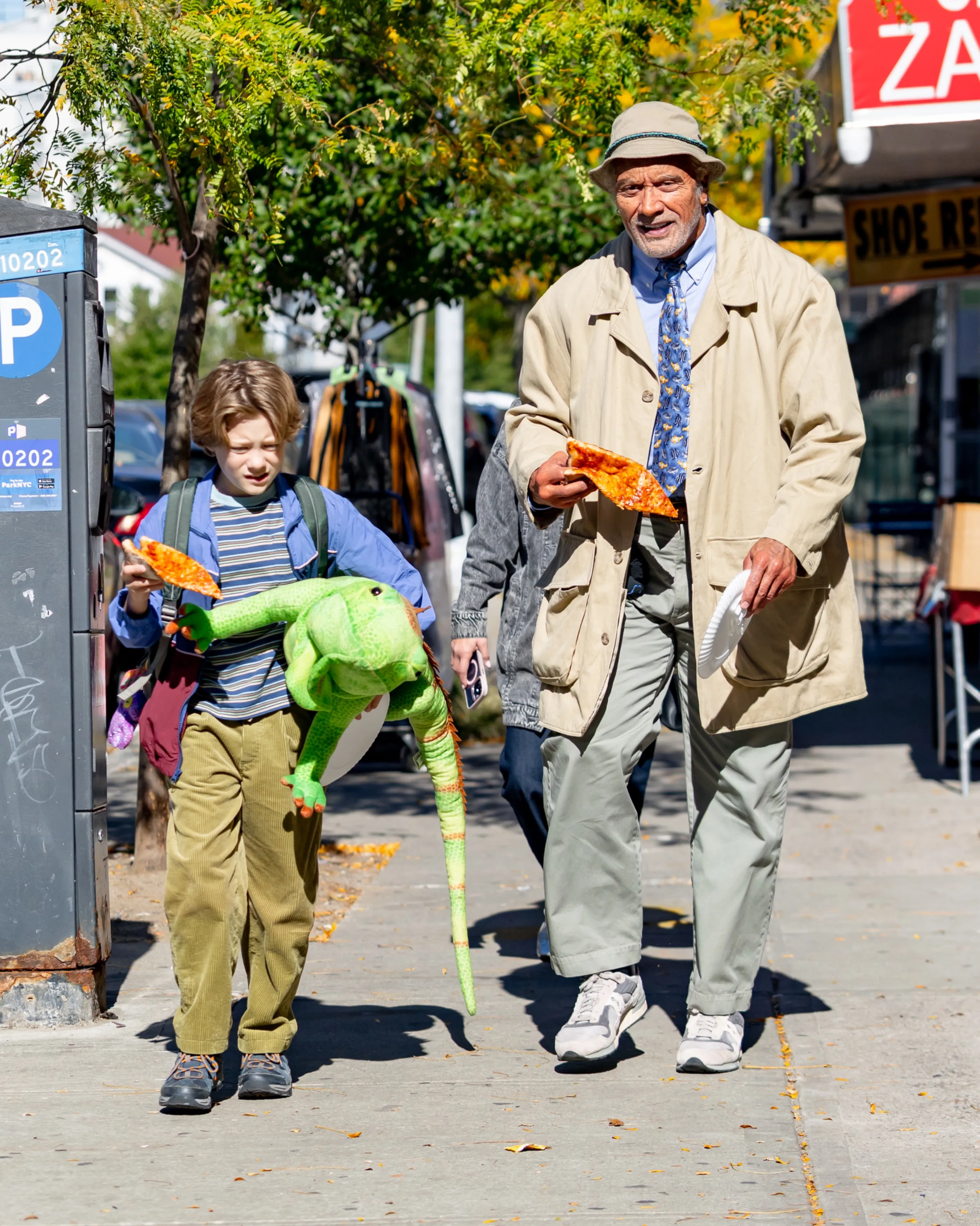 Dwayne Johnson, nearly unrecognizable in a tan trench coat, bucket hat, and beard, walks down a New York City street eating pizza alongside co-star Theo Clark Leber, who carries a large green lizard puppet and a slice of pizza.