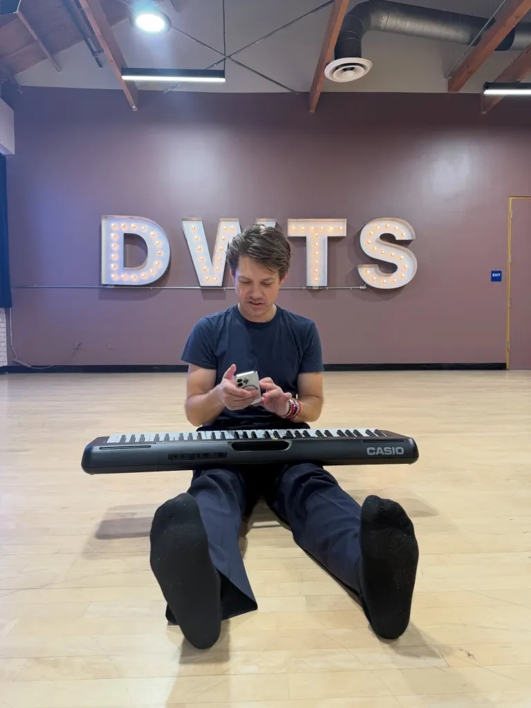 Taylor Hanson sits on a dance studio floor with a Casio keyboard in his lap, looking at his phone in front of a