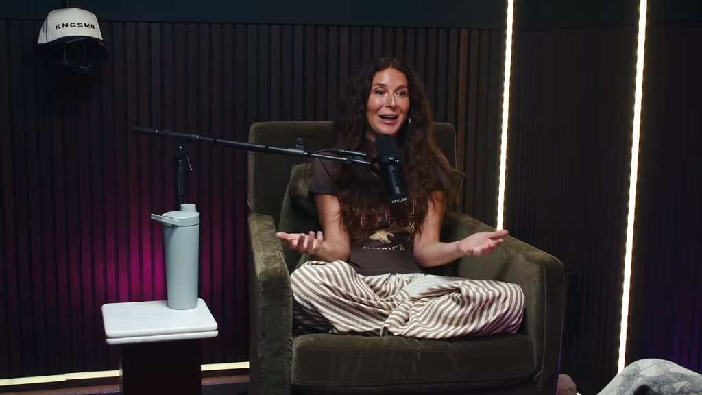 A woman with long brown hair, sitting cross-legged in a green armchair, gestures while speaking into a microphone in a studio setting.