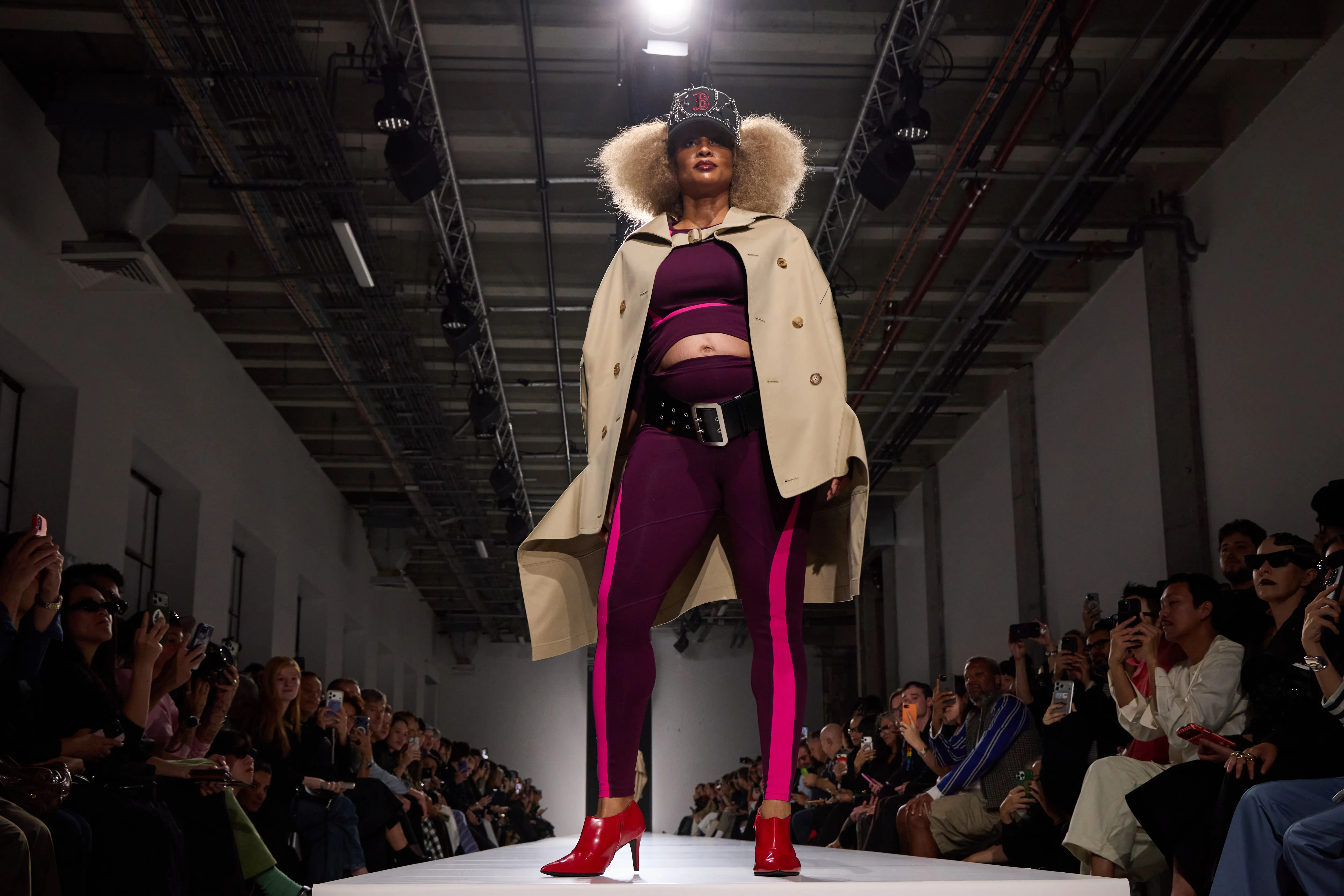 Ciara walks the runway during the Junya Watanabe show, wearing a tan trench coat, purple leggings with pink stripes, a baseball cap, and red heels.
