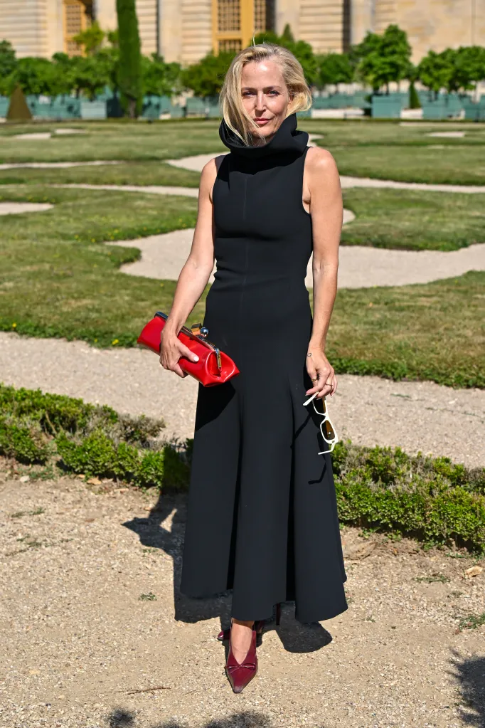 Gillian Anderson poses in a black dress holding a red clutch outdoors.