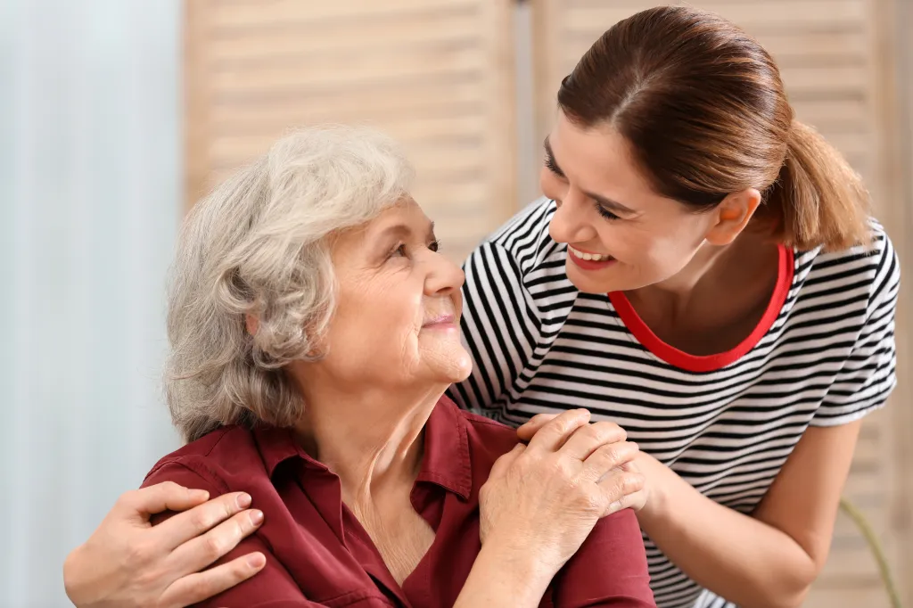 An elderly woman and her female caregiver smiling and looking at each other at home.