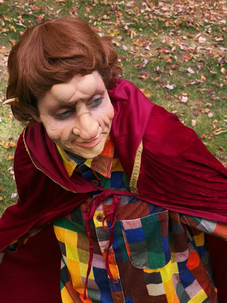 Elizabeth Gillies in a Halloween costume resembling a hobbit with prosthetic forehead and nose makeup, a reddish-brown wig with pointed ears, a patchwork button-down shirt, and a maroon velvet cloak.