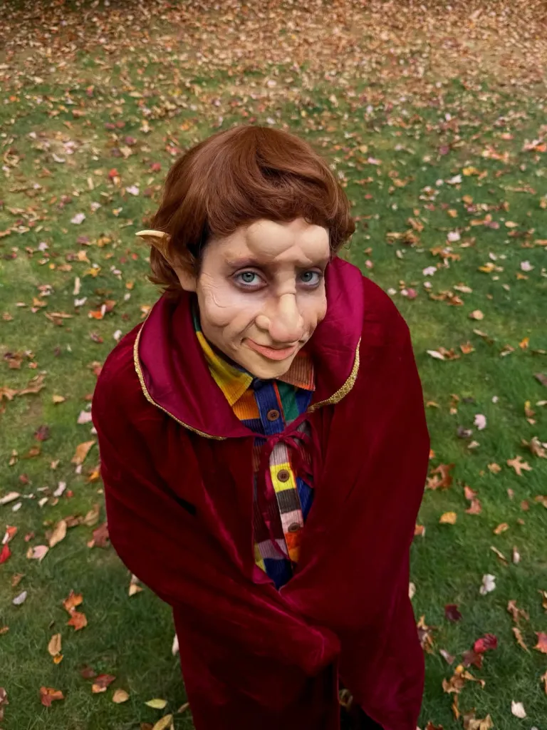 Elizabeth Gillies in a Halloween costume resembling a goblin, wearing a red cloak over a colorful plaid shirt, with prosthetics on her face and ears.