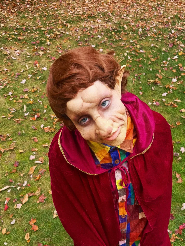 Elizabeth Gillies in a gnome costume, wearing a red hooded cape, a patterned shirt, and prosthetics for a gnome-like face.