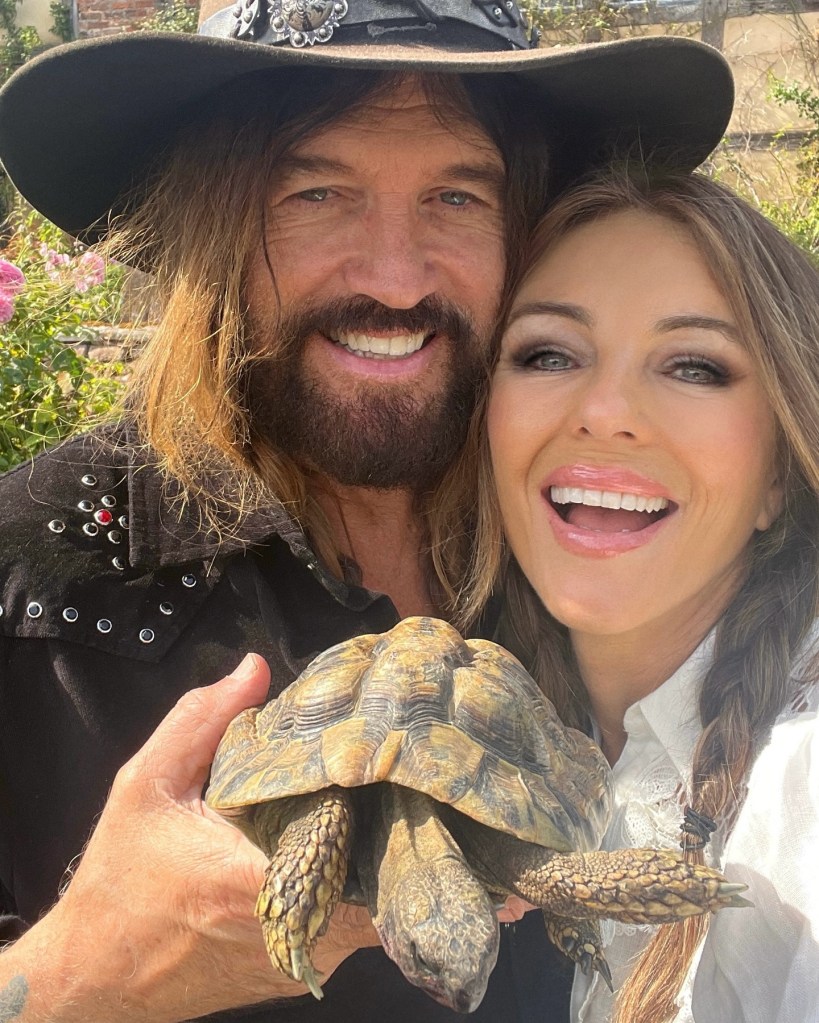 Elizabeth Hurley and Billy Ray Cyrus smiling together while holding a tortoise.