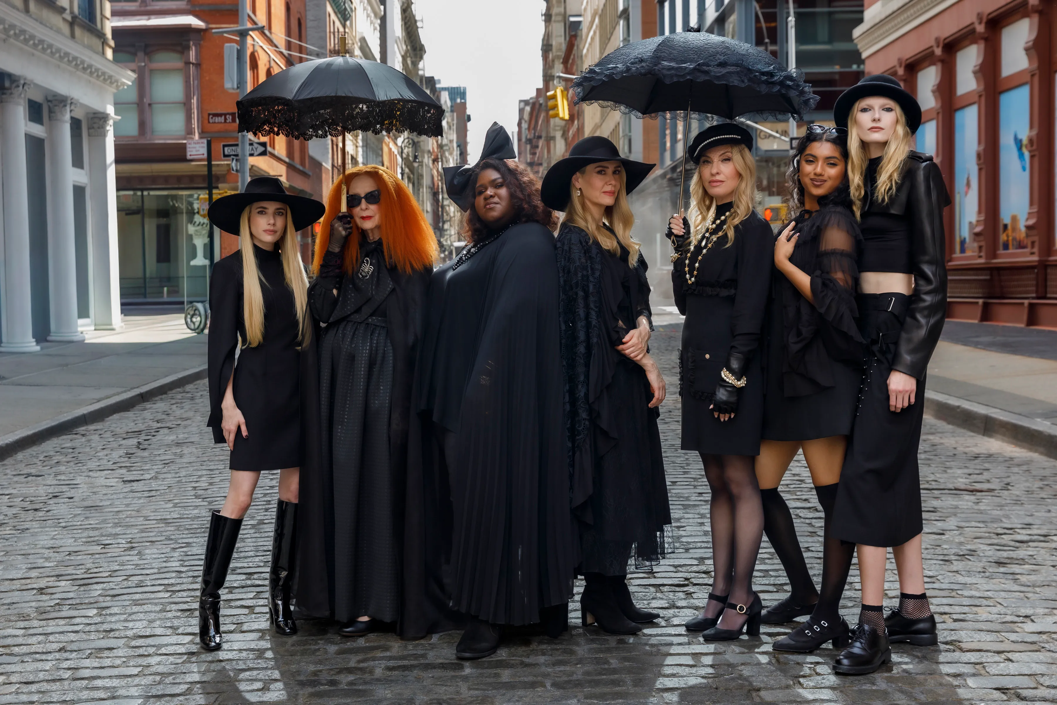 Cast members of American Horror Story, dressed in black and holding umbrellas, stand on a cobblestone street.