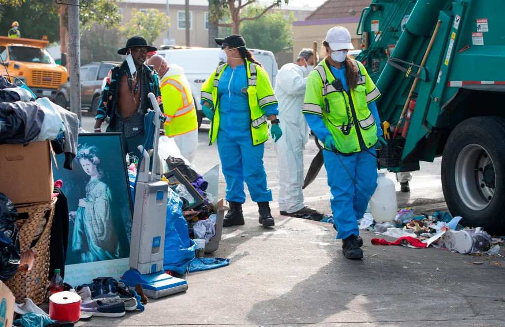 A sanitation worker in blue scrubs and a safety vest sprays chemicals on the street next to a garbage truck, while an unhoused man and activists watch nearby.