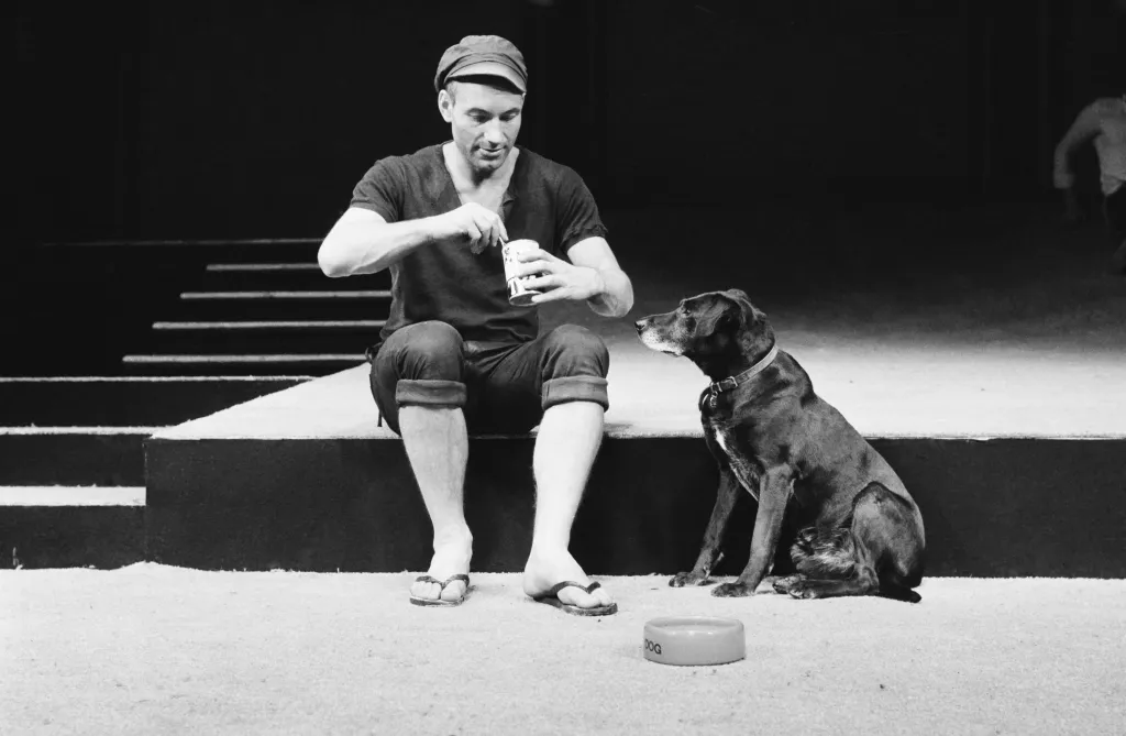 Black and white photo of actor Patrick Stewart as Launce, sitting on a stage and opening a can of food for his dog Crab.